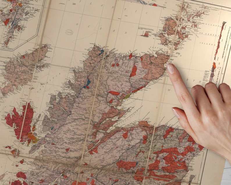 May include: A vintage map of Scotland with a finger pointing to the location of the Isle of Skye. The map is printed on paper with a cream background and features red, blue, and brown lines and markings.