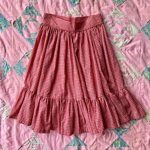 May include: A red and white gingham skirt with a ruffled hem. The skirt is made of a lightweight fabric and has a high waist. The skirt is displayed on a pink quilt.