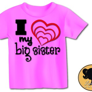 Puede incluir: Camiseta rosa con un gráfico negro que dice "Amo a mi hermana mayor" y un diseño de corazón rojo.