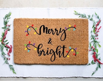 Puede incluir: Una alfombra de coco marrón con el texto "Merry & bright" en escritura negra. El texto está rodeado de luces de cadena de colores.