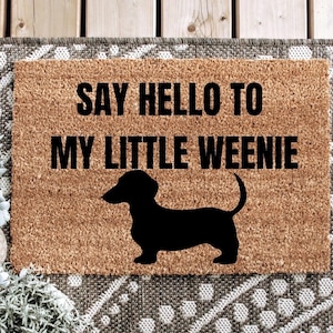 May include: A brown coir doormat with the text "Say hello to my little weenie" in black lettering. A black silhouette of a dachshund is below the text.