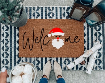 May include: A brown doormat with the word "Welcome" written in black script with a Santa Claus face in the middle of the word. The doormat is on a black and white patterned rug.