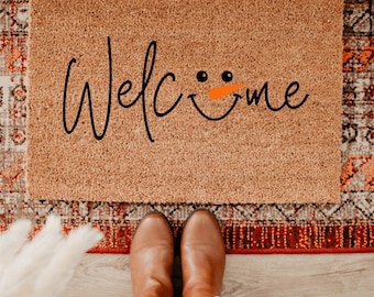 May include: A brown coir doormat with the word "Welcome" written in black cursive script. A snowman face with orange carrot nose is incorporated into the word "Welcome".