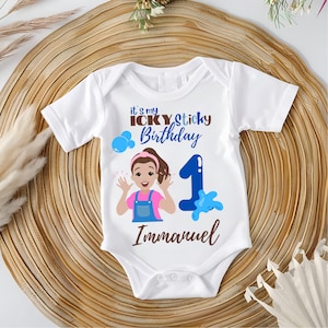May include: White baby onesie with a colorful graphic for a first birthday. The design includes the text "It's my Icky Sticky Birthday", a cartoon illustration of a girl, the number 1, and the name "Immanuel".