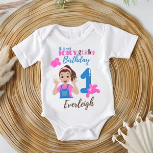 May include: White baby onesie with a colorful graphic. The design includes the text "It's my Icky Sticky Birthday" and the number "1". A cartoon girl and the name "Everleigh" are also featured.
