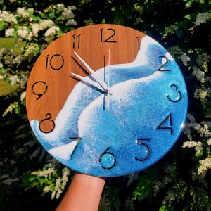 May include: Round wooden wall clock with a beach-themed design. The clock face features a brown wooden center with black numerals and silver hands. The bottom half is a blue and white resin wave design.