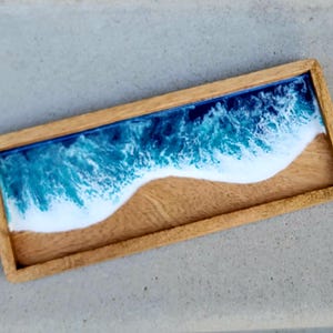 May include: A wooden tray with a blue and white resin ocean wave design. The wave is breaking over a sandy beach.