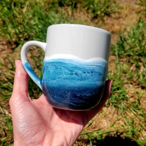 May include: A white ceramic mug with a blue and teal ocean wave design. The mug has a rounded shape and a handle.
