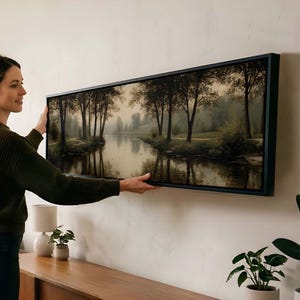 Puede incluir: Una pieza de arte mural panor&aacute;mico que presenta una escena fluvial serena con &aacute;rboles y reflejos, encerrada en un marco negro fino. La obra muestra una paleta de colores terrosos y apagados con una iluminaci&oacute;n suave y calmada.
