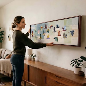 May include: A long, rectangular wall art piece featuring a flock of colorful, stylized birds in flight against a soft, segmented background of pale blue, green, and cream tones, all enclosed within a dark wood frame.