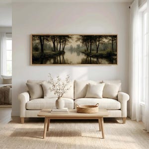 Puede incluir: Un cuadro panor&aacute;mico de paisaje en un marco de madera que representa un r&iacute;o tranquilo bordeado de &aacute;rboles oscuros, expuesto sobre un sof&aacute; de rayas beige con cojines claros, una mesa de centro de madera, un jarr&oacute;n blanco y una bandeja trenzada.