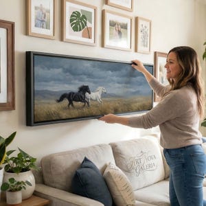 May include: A panoramic landscape painting in a black frame featuring a black horse and a white horse galloping across a golden grassy field under a dark, cloudy sky. The artwork is displayed horizontally on a wall above a beige sofa.