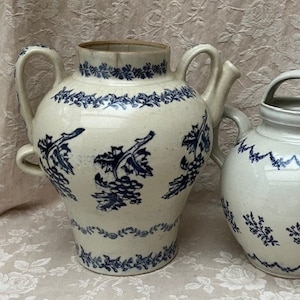 Puede incluir: Dos teteras antiguas de cerámica blanca con diseños florales azules. La tetera más grande tiene dos asas y un pico, mientras que la más pequeña tiene un asa. Ambas tienen adornos azules.