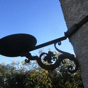 May include: A wrought iron wall-mounted bird feeder with a circular tray and decorative scrollwork. The feeder is painted black and is attached to a textured stucco wall. The background features a clear blue sky and green foliage.