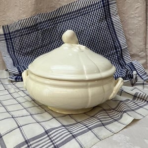 May include: Cream-coloured, lidded soup tureen with side handles. The tureen sits on a white and blue plaid cloth. The lid has a decorative knob. The tureen is a classic piece for serving soups and stews.