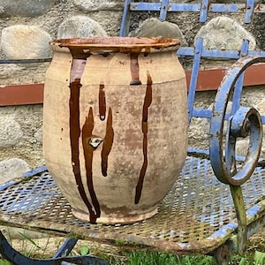 May include: A cylindrical earthenware pot with a wide opening and a tapered base. The exterior is a light tan color with dark brown glaze drips running vertically from the rim down the sides of the vessel, creating a textured appearance.