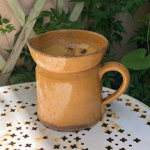 May include: A mustard yellow ceramic pitcher with a wide, flared rim and a single side handle. The exterior features a smooth, glazed finish with a visible crack near the top rim and a darker, weathered base. The interior is also glazed.