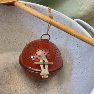 May include: A vintage red tea infuser with a perforated spherical design, suspended by twine from a wooden spoon. The infuser features a metal clasp and is set against a speckled white enamel bowl.