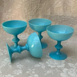 May include: A set of three bright turquoise glass sherbet cups, each featuring a wide, shallow bowl and a pedestal base with a distinct central ring detail. Two cups stand upright, while one cup is positioned on its side in the foreground.