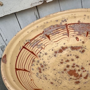 May include: A large, round, cream-colored ceramic bowl featuring a wide, flared rim. The interior surface is decorated with a hand-painted, reddish-brown marbled pattern of concentric lines and scattered, irregular spots across the base.