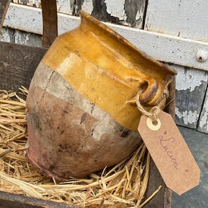 May include: A rustic earthenware pot with a glazed yellow upper section and a natural terracotta base. The pot features two side handles and a brown paper tag tied with twine, which has the name Lucien written in cursive script on the front.