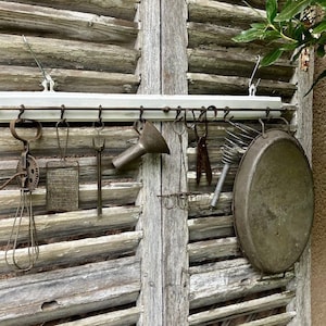 May include: A white-painted kitchen utensil rack with various antique cooking tools hanging from it. The tools include a whisk, grater, funnel, and a large pan. The rack is mounted on weathered wooden shutters.