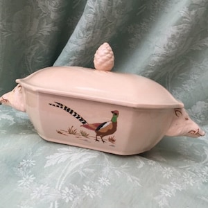 Puede incluir: Una fuente de cerámica rosa claro con tapa y asas con forma de cabeza de cerdo. La fuente presenta una ilustración de faisán y un asa en forma de piña. Un artículo vintage.