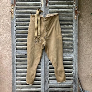 May include: High-waisted tan trousers featuring a button-up fly, a high waistband with multiple buttons for attachment, and a pair of matching tan suspenders with metal clips. The pants have a tapered leg design and a structured, tailored appearance.