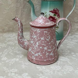 Peut inclure: Une théière en émail moucheté rose et blanc avec un couvercle assorti, un bec verseur courbé et une anse, placée devant une cafetière en émail bleu clair et blanc plus haute, ornée d'un motif floral de roses roses et de feuilles vertes.