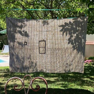 May include: A striped fabric hanging outdoors, featuring vertical black and beige stripes. The fabric has two black geometric shapes. The scene is set in a garden with a pool and a chair visible in the background.
