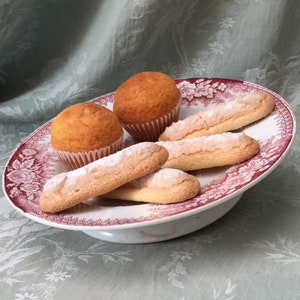 Könnte beinhalten: Zwei goldbraune Muffins in Papierförmchen und drei längliche Löffelbiskuits mit Zucker bestäubt liegen auf einem weißen Keramikteller, der am Rand mit einem roten Blumenmuster verziert ist.