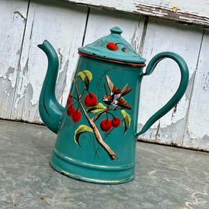 Puede incluir: Una cafetera de esmalte color verde azulado con un diseño pintado a mano de cerezas rojas brillantes sobre una rama marrón con hojas verdes y un pequeño pájaro marrón. La cafetera tiene una tapa a juego, un asa curva y un pico largo.