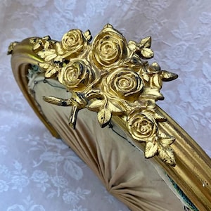 Könnte beinhalten: Ein goldfarbener, verzierter Bilderrahmen mit einem Rosenstrauß oben. Die Rosen und Blätter sind detailliert und verleihen eine dekorative Note. Der Rahmen hat eine geschwungene Form und einen weichen, neutralfarbenen Stoffrücken.