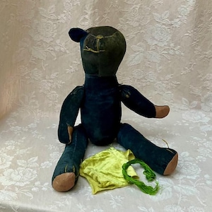 Puede incluir: Un oso de peluche de terciopelo azul oscuro con costuras visibles en la cabeza y detalles de tela color canela en las orejas, manos y pies. Junto al oso hay un pequeño trozo de tela amarilla brillante doblada y un hilo de bordar verde.