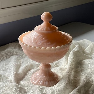Może przedstawiać: Wazon na cukierki z różowego szkła w stylu vintage z pokrywką. Naczynie ma podstawę na cokole i falisty brzeg. Pokrywka zwieńczona jest małym, okrągłym guzikiem. Naczynie zdobione jest wytłoczonymi wzorami kwiatowymi i stoi na białej koronkowej tkaninie.