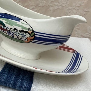 May include: White ceramic gravy boat and saucer set. The boat has a spout and handle. The set is decorated with blue stripes and a landscape scene. The saucer has blue and red stripes.