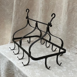 Puede incluir: Un estante negro de hierro forjado para ollas con base rectangular y soportes curvos. Cuenta con múltiples ganchos para colgar utensilios de cocina y dos ganchos grandes en la parte superior para el montaje. El estante se exhibe sobre un mantel con estampado floral.