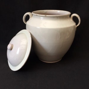 Op de afbeelding: Een crèmekleurige keramische pot met een deksel, met twee lusvormige handvatten. Het deksel ligt opzij en onthult de binnenkant. De pot heeft een afgeronde body en een licht uitlopende rand. De achtergrond is zwart.