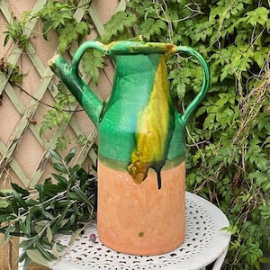 Puede incluir: Una jarra de cerámica hecha a mano con un diseño único. La jarra presenta una combinación de colores vibrantes, con una parte superior esmaltada en verde y amarillo y una base de color terracota. Tiene un pico y dos asas. La jarra está sobre una mesa blanca.