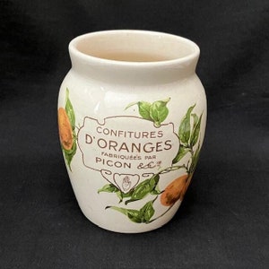 Könnte beinhalten: Ein cremefarbener Keramiktopf mit einem dekorativen Etikett mit der Aufschrift CONFITURES D'ORANGES FABRIQUÉES PAR PICON. Der Topf ist auf beiden Seiten des zentralen Textetiketts mit Illustrationen von Orangenfrüchten und grünen Blättern verziert.