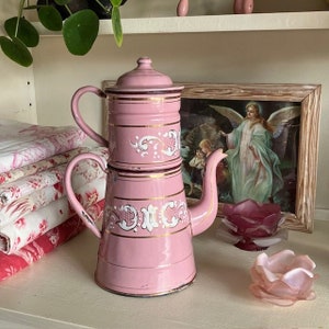 Könnte beinhalten: Eine rosa Vintage-Kaffeekanne mit Deckel, Griff und Ausguss. Die Kanne hat goldfarbene Verzierungen und weiße dekorative Akzente. Sie steht auf einem weißen Regal mit einem gerahmten Bild und zwei dekorativen Glasschalen.