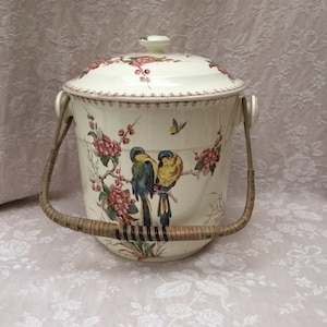 May include: Cream-colored ceramic ice bucket with a lid, featuring a floral and bird design. The bucket has a woven handle and is decorated with pink flowers, green leaves, and two colorful birds. A butterfly is also depicted.