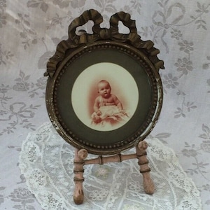 Puede incluir: Marco de fotos ovalado antiguo con un retrato de bebé en tonos sepia. El marco es de color bronce con una cinta decorativa en la parte superior y un pequeño soporte. El bebé está sentado sobre una superficie blanca.