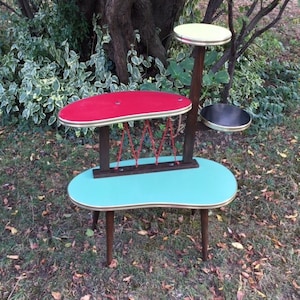 Op de afbeelding: Een vintage plantenstandaard met een turquoise ovale basis, een rode ovale bovenkant en een gele cirkelvormige bovenkant. De standaard is gemaakt van hout en heeft een uniek ontwerp. De hoogte is ongeveer 75 cm.