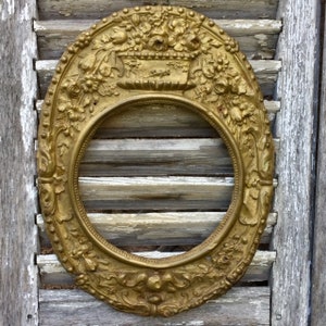 Pode incluir: Uma moldura oval de cor dourada com entalhes em relevo intrincados de arranjos florais e um motivo central de cesta. A moldura possui uma borda interna com contas e um design decorativo de volutas ao longo da borda inferior, criando textura.