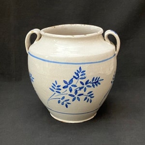 Op de afbeelding: Een crèmekleurige keramische pot met twee handvatten en blauwe bloemmotieven. De pot heeft een blauwe band in het midden en aan de basis. Het ontwerp is rustiek en vintage.