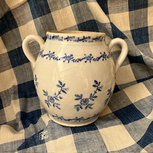 May include: A white ceramic jar with two handles, decorated with blue floral patterns. The jar has a rounded body and a slightly flared rim. The blue floral design includes sprigs and garlands, adding a touch of elegance to this decorative piece.