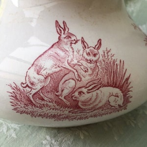 Könnte beinhalten: Weißer Keramikkrug mit einer roten Illustration von drei Hasen in einem grasbewachsenen Feld. Die Hasen sind in verschiedenen Posen dargestellt. Das Design ist klassisch und im Vintage-Stil gehalten.