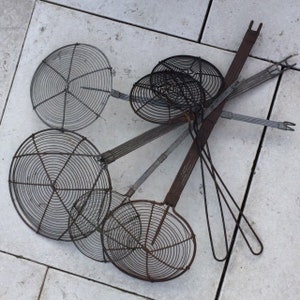 May include: A collection of vintage metal skimmers and strainers with wire mesh bowls and long handles. The kitchen tools are in various sizes and show signs of age and use, with a rustic, weathered appearance. The arrangement is on a light-colored, textured surface.