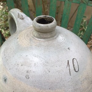 May include: A light gray stoneware jug with a rounded body, a single side handle, and a narrow neck opening. The surface features a weathered texture with visible imperfections and the number 10 clearly marked in a dark, handwritten-style font.